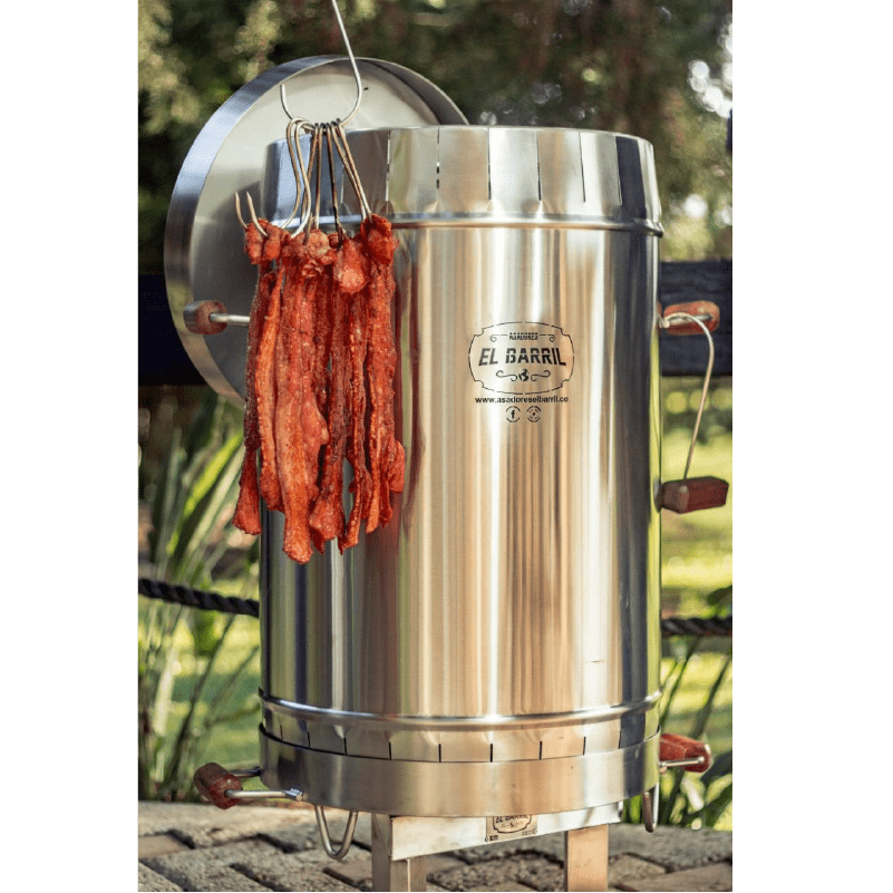 Barril asador mediano con tapa lateral y tiras de carne adobada colgadas de los ganchos, en un jardín al aire libre