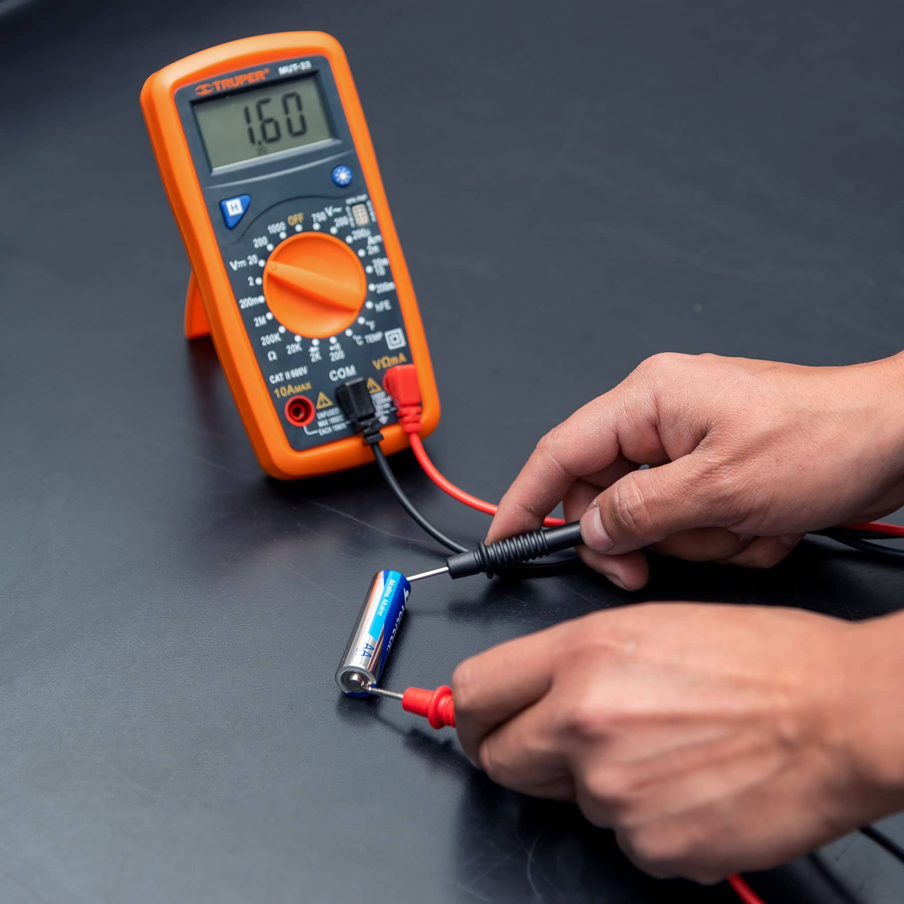 Polímetro digital Truper MUT-33 midiendo el voltaje de una pila AA con las puntas de prueba sobre una mesa de trabajo