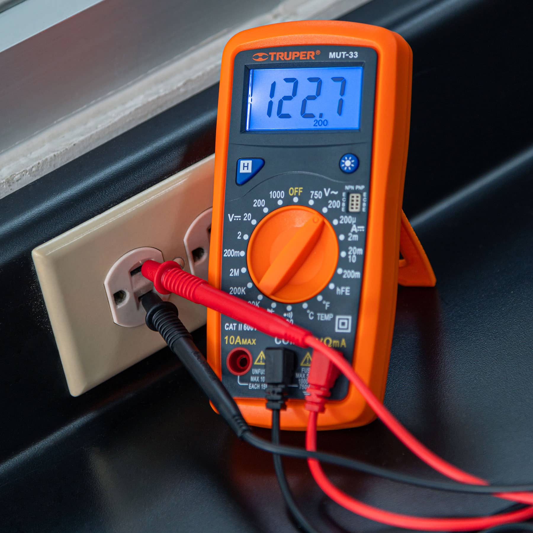Polímetro digital Truper MUT-33 con pantalla iluminada midiendo el voltaje de un tomacorriente en una instalación