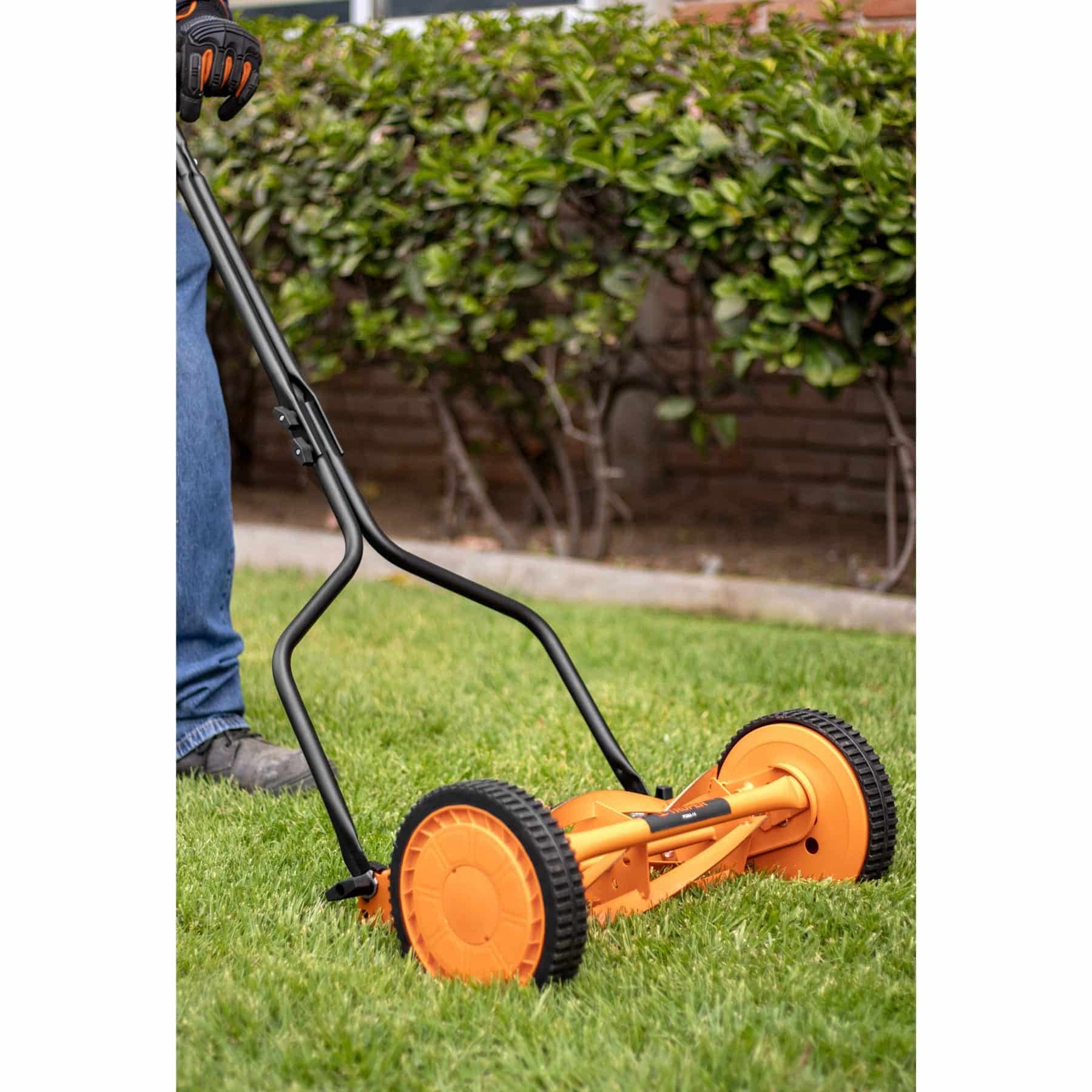 Podadora manual Truper POMA-14 cortando césped en un jardín con cerca viva al fondo, operada por persona con guantes
